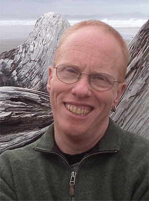 Head shot of a man with pale skin, short reddish hair and glasses, and an earring in one ear. Behind him is some driftwood.
