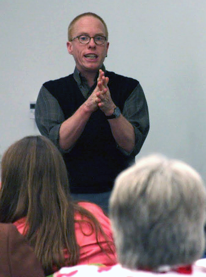 Eli stands speaking in front of two people who are sitting.