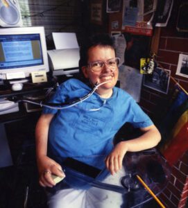 A white woman with short dark hair sits in a wheelchair in front of a computer. She has a breathing tube in her nose.