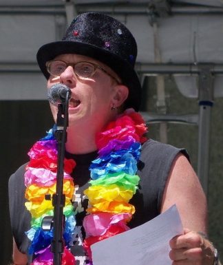 Eli stands speaking into a mic. He's wearing a black hat and a rainbow colored lei.