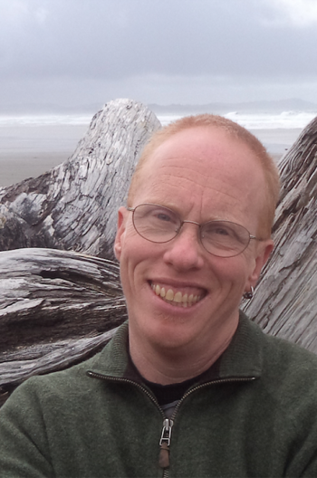 Head shot of a man with pale skin, short reddish hair and glasses, and an earring in one ear. Behind him is some driftwood.